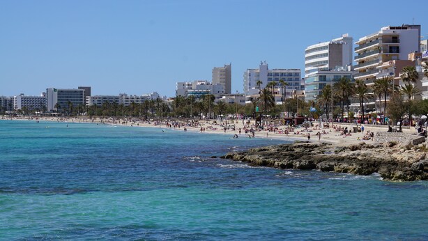 Una playa con agua azul cristalina, orilla de arena y edificios al fondo.
