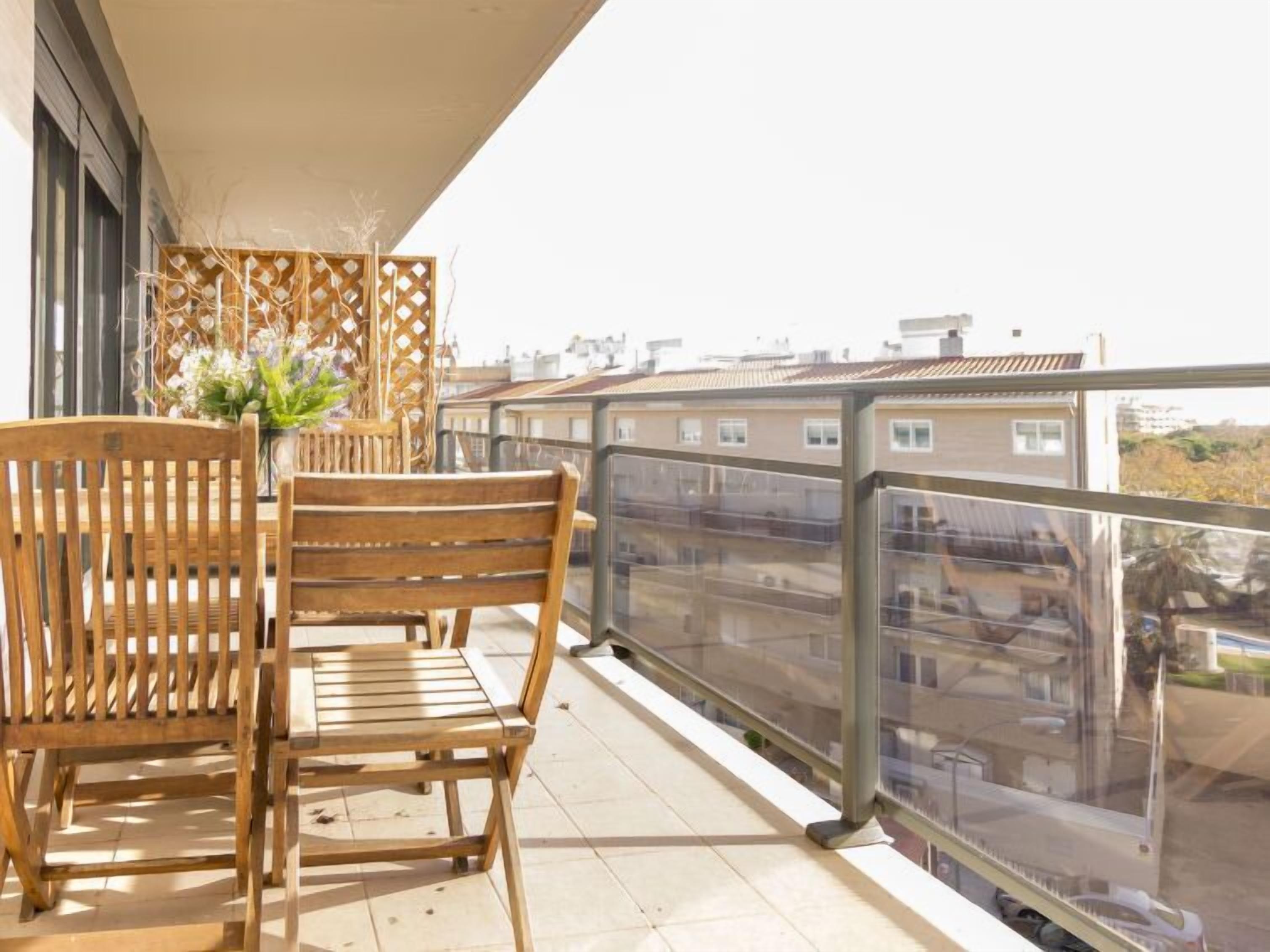 A balcony with two wooden chairs and a table, a decorative plant, and a view of buildings through a glass railing.