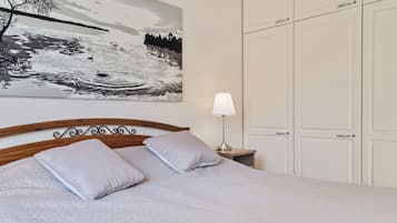A bedroom with a bed, pillows, a nightstand with a lamp, a painting on the wall, and a wardrobe.