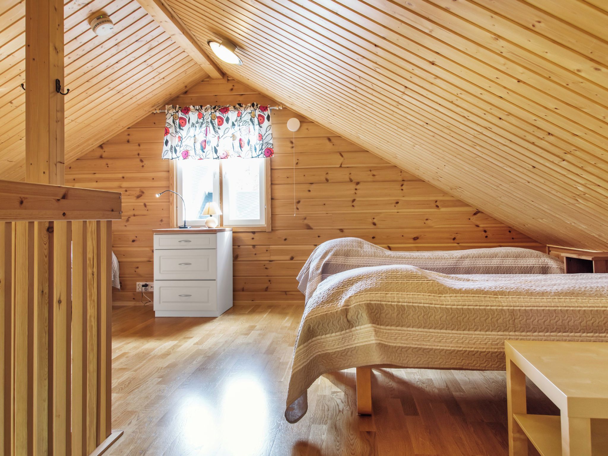 A cozy attic bedroom with wooden walls and sloped ceilings, featuring two beds, a dresser, and a window with floral curtains.
