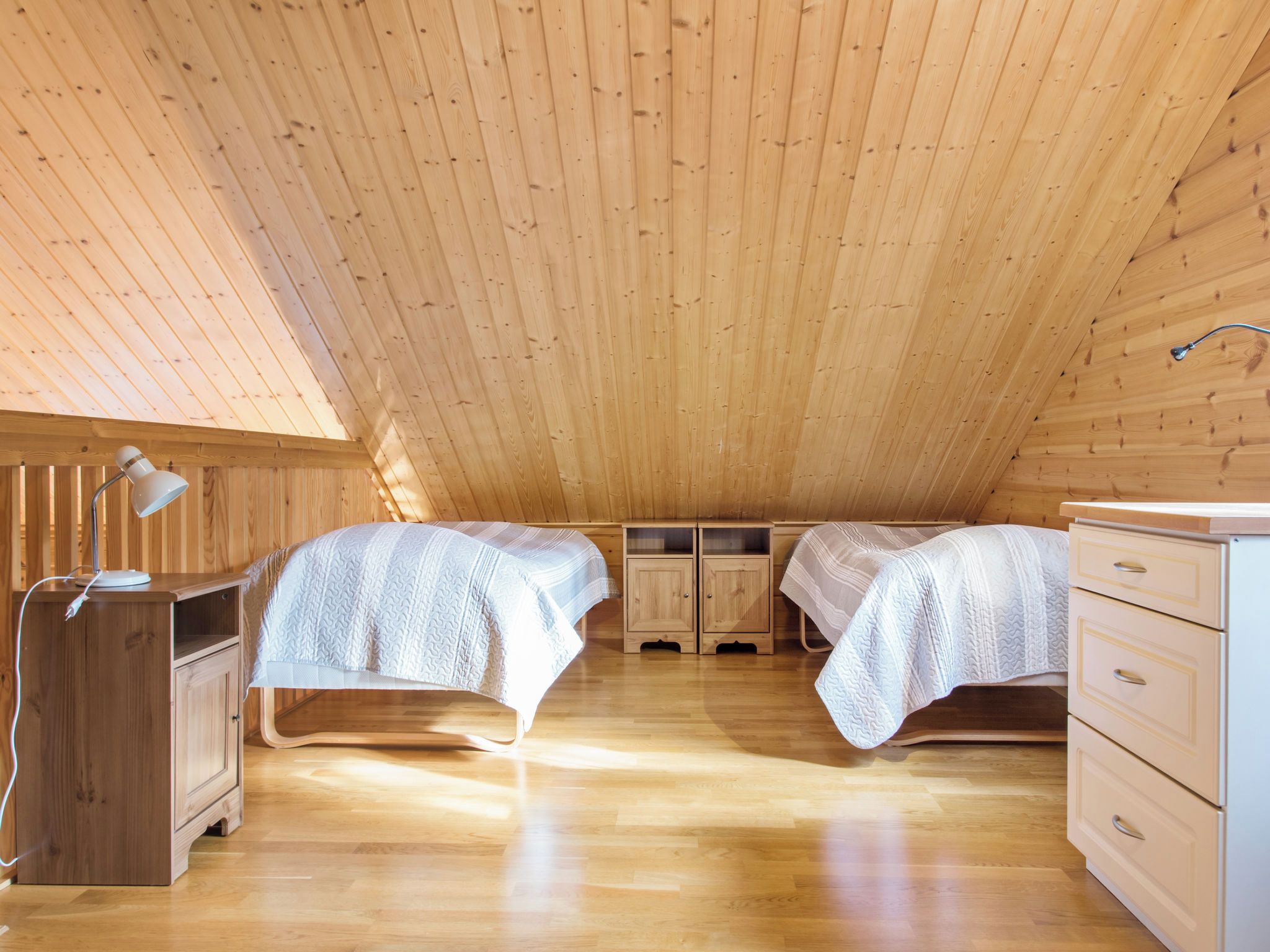 Une pièce avec un plafond en pente, deux lits avec du linge de lit blanc, un bureau avec une lampe et des armoires en bois.
