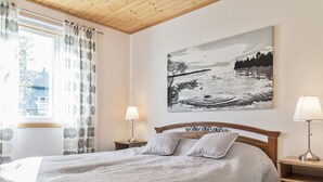 Quarto com cama, duas luminárias de cabeceira, painel de cabeceira de madeira, mesa de cabeceira, armário de madeira, janela com cortinas e uma pintura pendurada na parede.