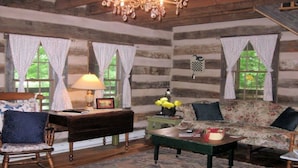 A rustic log cabin interior with a wooden bench, a green coffee table, a floral sofa, and a chandelier. - Tavern River Cabin, Historical,  Riverfront, Log, Hottub, Large Stone Fireplace (Luray)