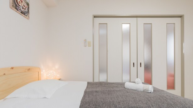 Una camera da letto con una testiera in legno, un letto con biancheria bianca, una coperta grigia e un asciugamano arrotolato. C'è una lampada a parete e un quadro incorniciato sopra il letto.