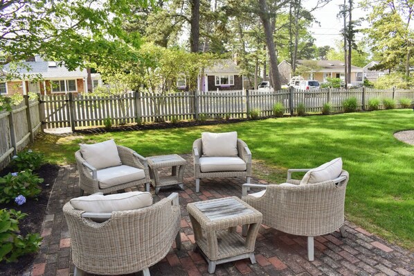 Relax in a serene outdoor seating area with wicker chairs and a small table on a brick patio surrounded by a lawn.