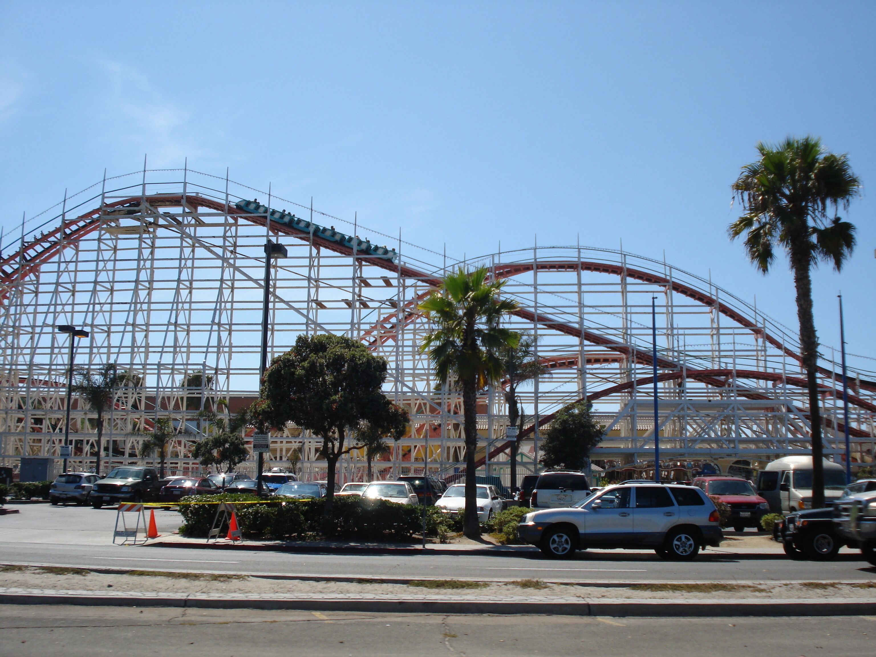 A large amusement park with a roller coaster under construction, surrounded by palm trees and parked cars.