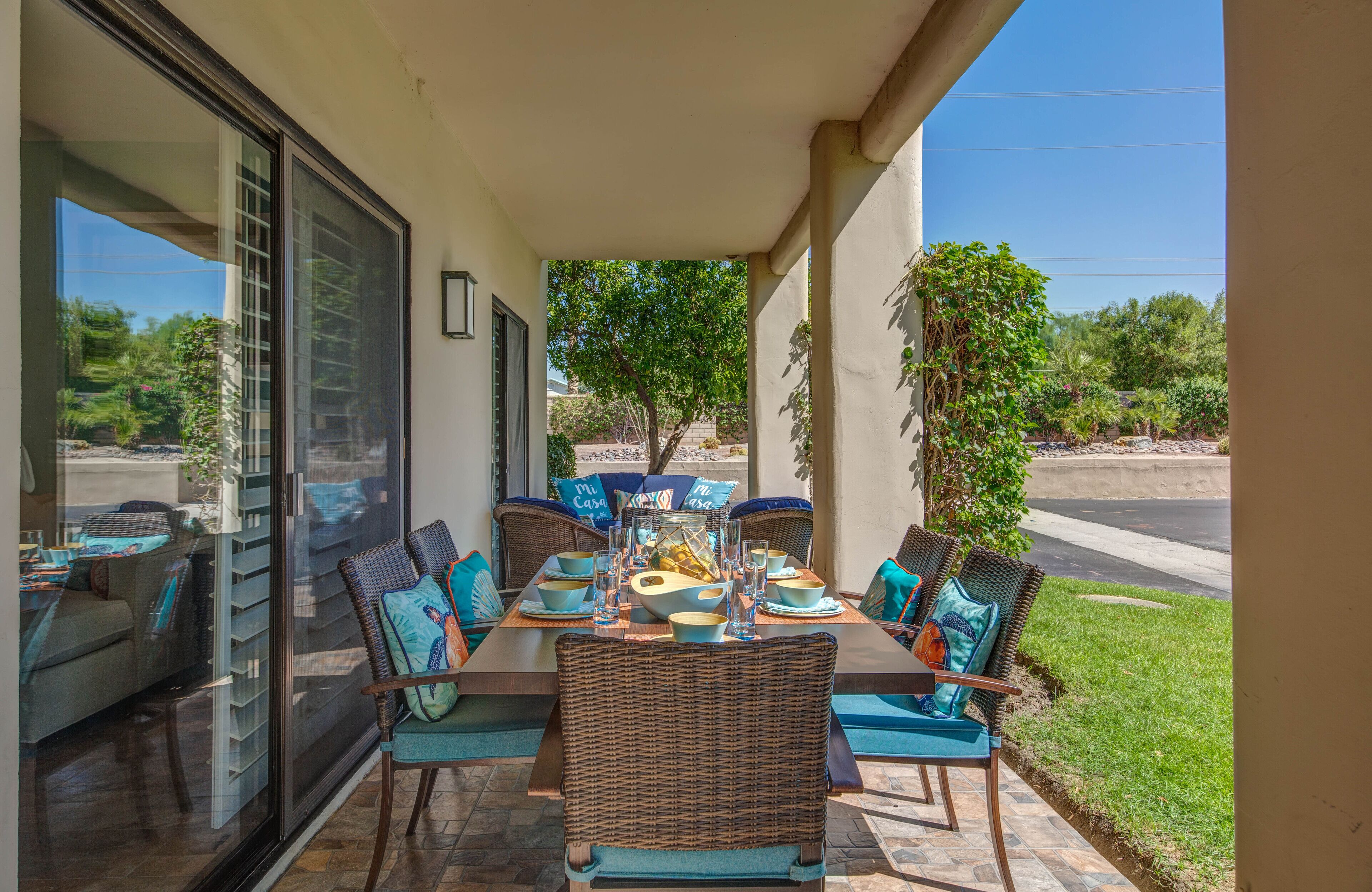 An outdoor dining area with wicker furniture and teal cushions, overlooking a landscaped garden and a road.