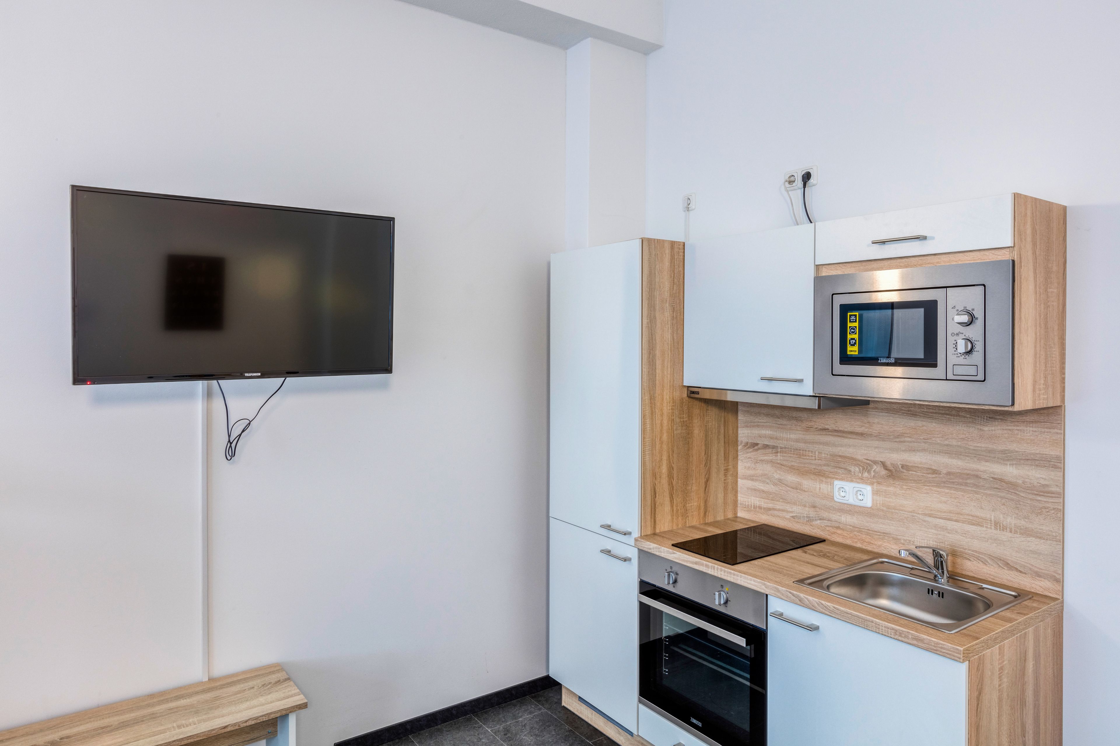 A compact kitchen with a built-in microwave, oven, and sink, a wooden countertop, and a flat-screen TV mounted on the wall.