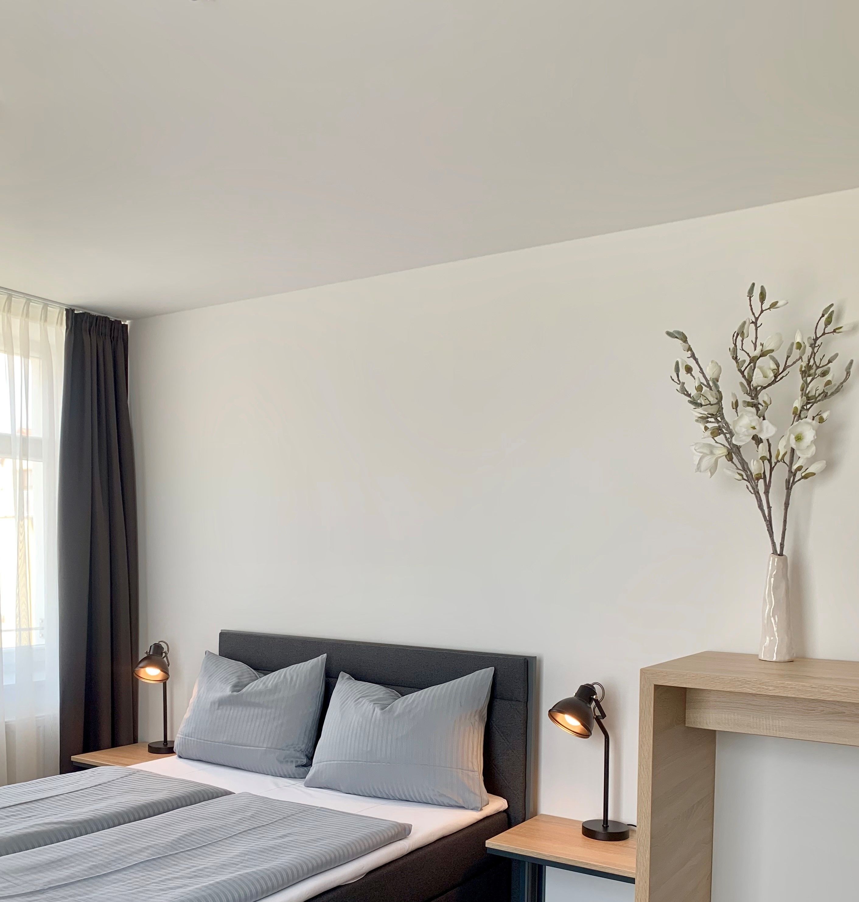 A modern bedroom with a bed, two bedside lamps, a nightstand, a vase with flowers, and a window with curtains.