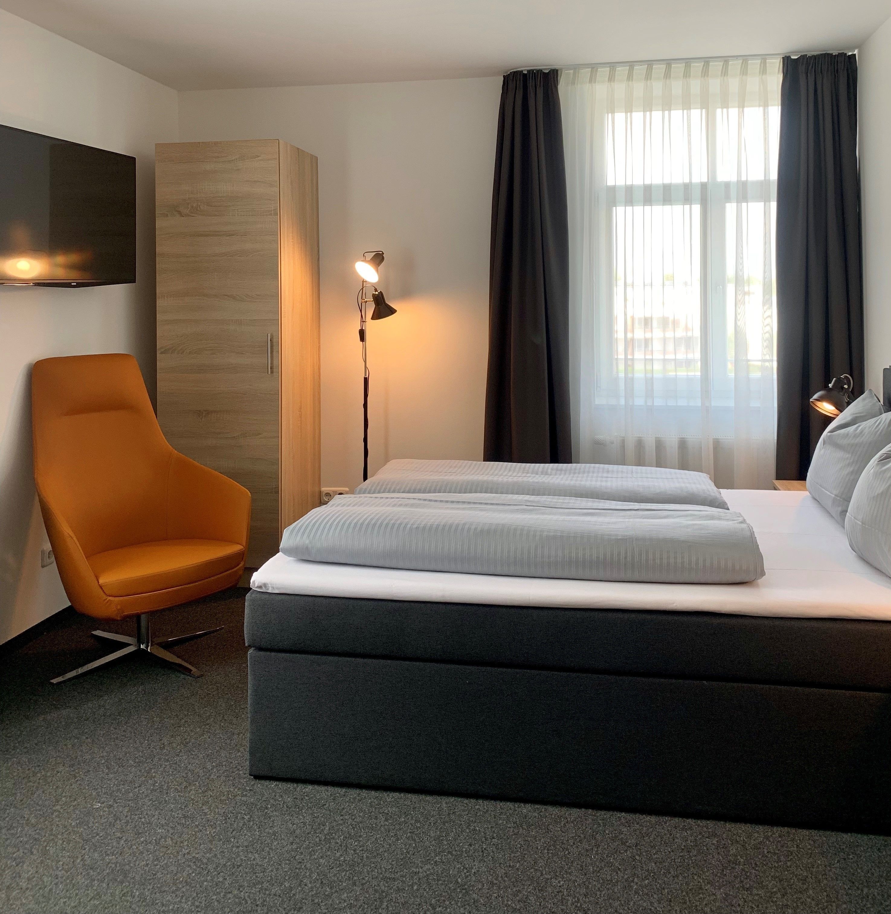 A modern hotel room with a large bed, a chair, a TV, and a window with curtains.