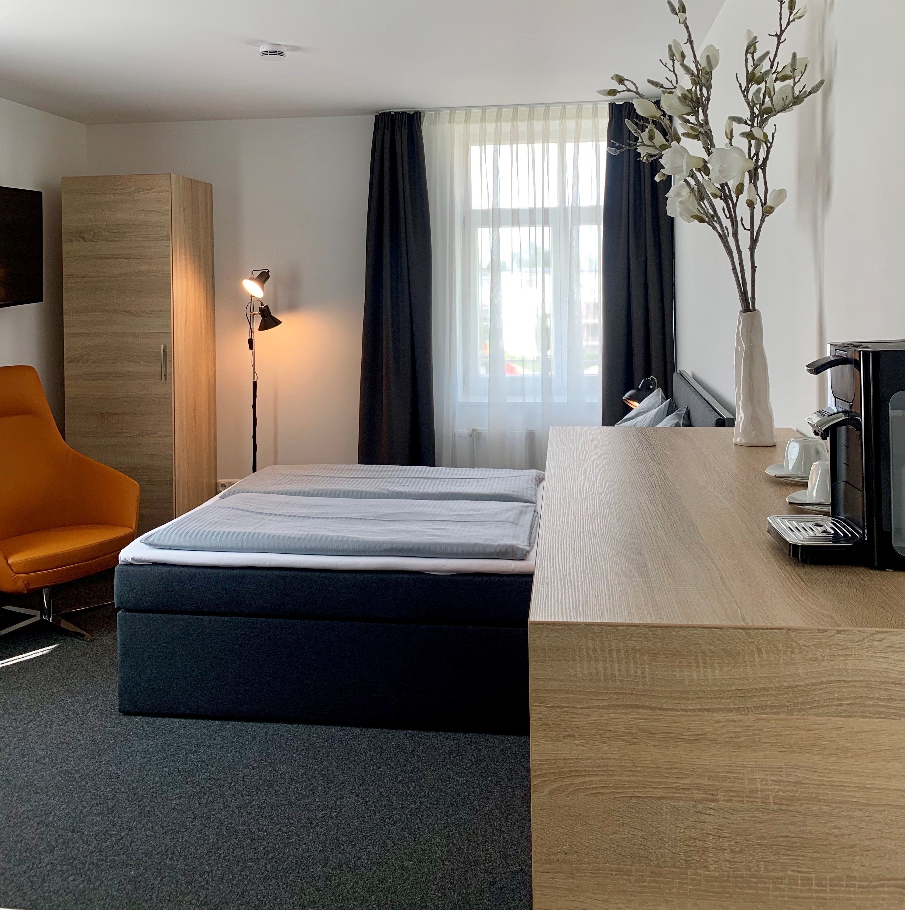 Une chambre d’hôtel comprenant un lit, un bureau avec une machine à café, une chaise, une armoire et une fenêtre avec des rideaux.