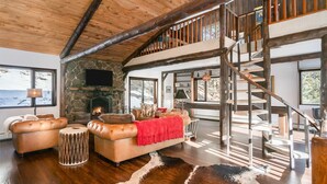 A spacious living room with a stone fireplace, leather sofas, a cowhide rug, and a wooden staircase leading to an upper level. - Luxury Mountain Retreat Near Estes Park Stunning Views, Perfect Family Getaway (Allenspark)