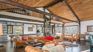 A spacious living room with leather sofas, a wooden staircase, and large windows offering views of the outdoors. - Luxury Mountain Retreat Near Estes Park Stunning Views, Perfect Family Getaway (Allenspark)