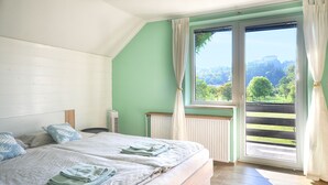 A bedroom with two beds, a radiator, a window with curtains, and a view of greenery and a building.