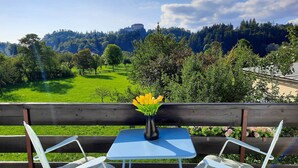 A wooden bench with a table and chairs, a vase of yellow flowers, and a view of a green field and forested hills.