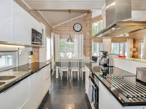 Cozinha moderna com armários brancos, bancada preta e eletrodomésticos de aço inoxidável. Há uma área de jantar com cadeiras brancas e uma mesa, e um relógio na parede.