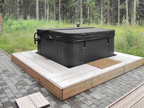 Un hidromasaje exterior negro sobre una plataforma de madera, con un bosque cubierto de hierba al fondo.