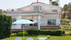 Une maison à deux étages, avec un toit en tuiles, un patio couvert comprenant des chaises longues et un parasol, ainsi qu’une pelouse bien entretenue.