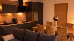 A modern living room with a dark grey sectional sofa, a dining area with wicker chairs, a kitchen with dark cabinetry, and a wooden door. - Charming village house (Santa Maria Figaniella)