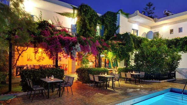An outdoor pool area with tables and chairs, surrounded by lush greenery and flowering plants. - Spacious apartment just 2 min walk from the beach! (Plakias)