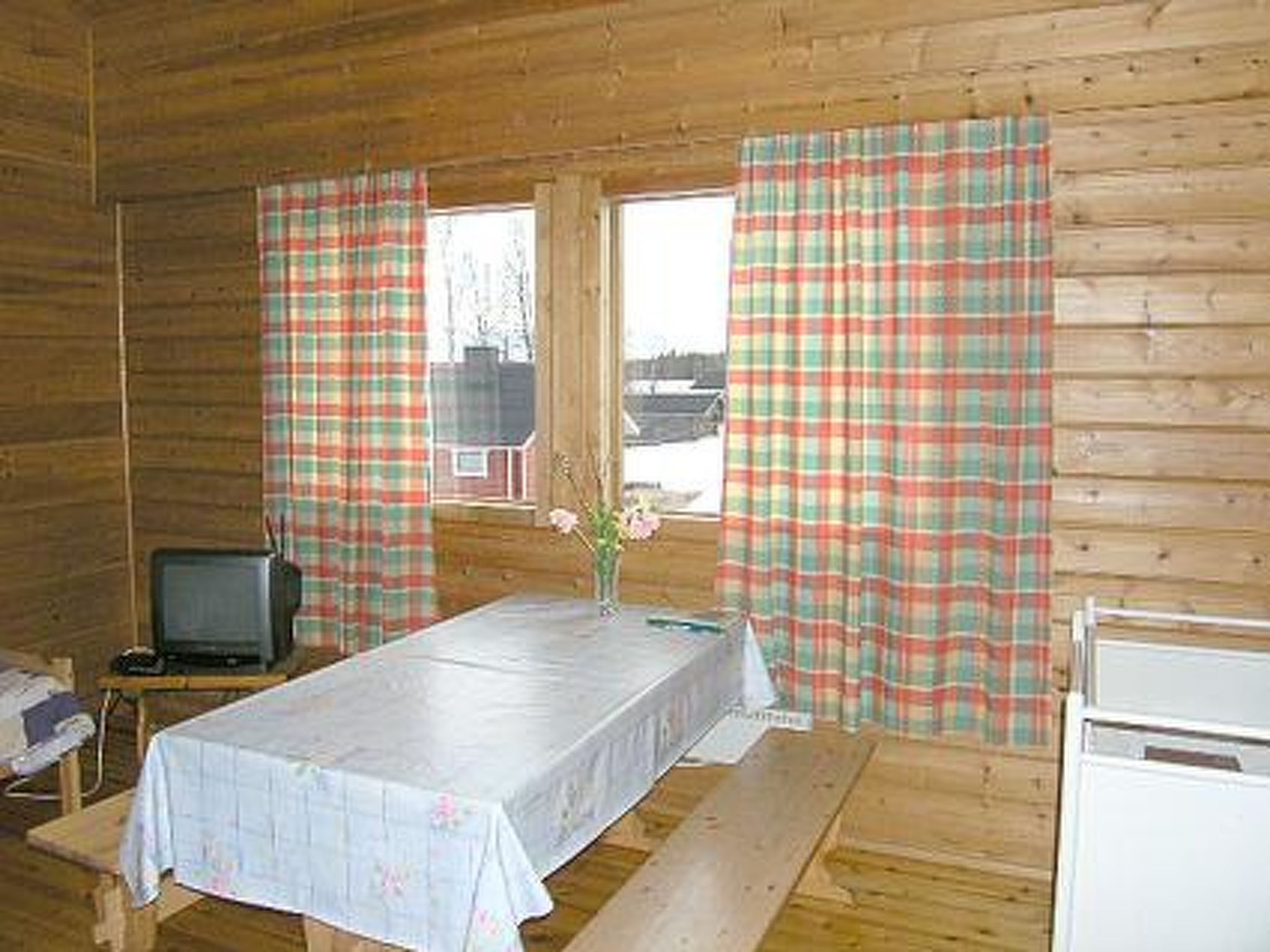 Een houten kamer met een tafel met tafelkleed, een televisie en geruite gordijnen.