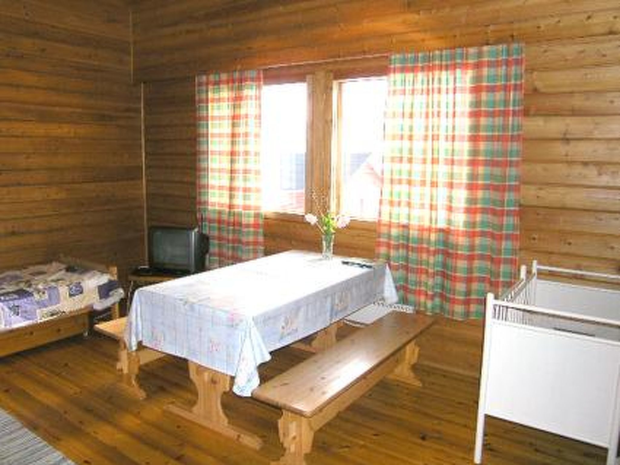 Een blokhut met houten inrichting, een bed, een tafel met tafelkleed, een bank, een televisie en geruite gordijnen.