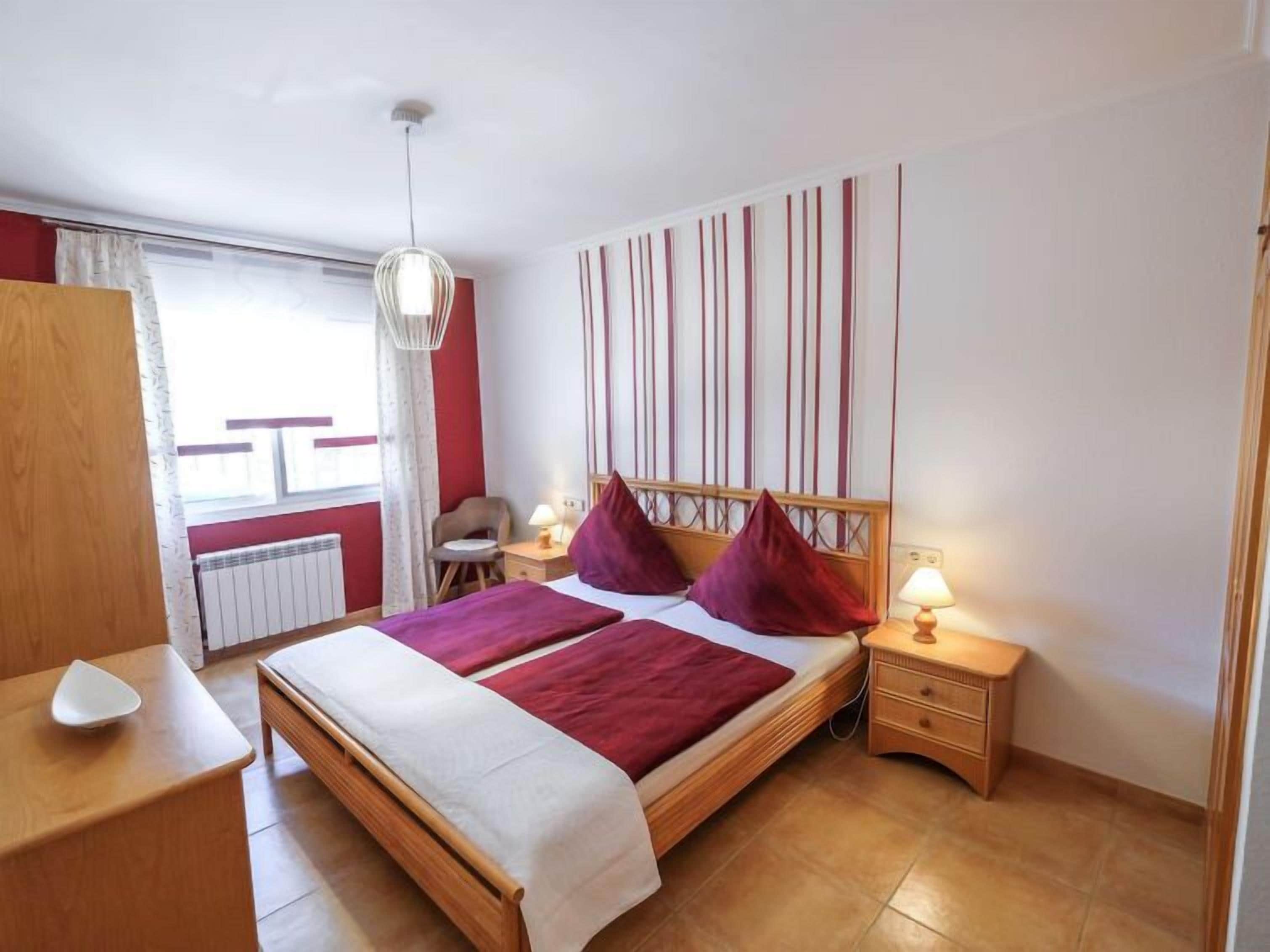 A bedroom with a wooden bed, a nightstand with a lamp, a chair, and a window with curtains.