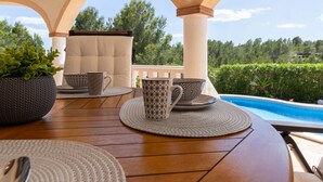 A wooden table with patterned tableware and a potted plant, set under a covered patio with a view of a swimming pool and greenery.