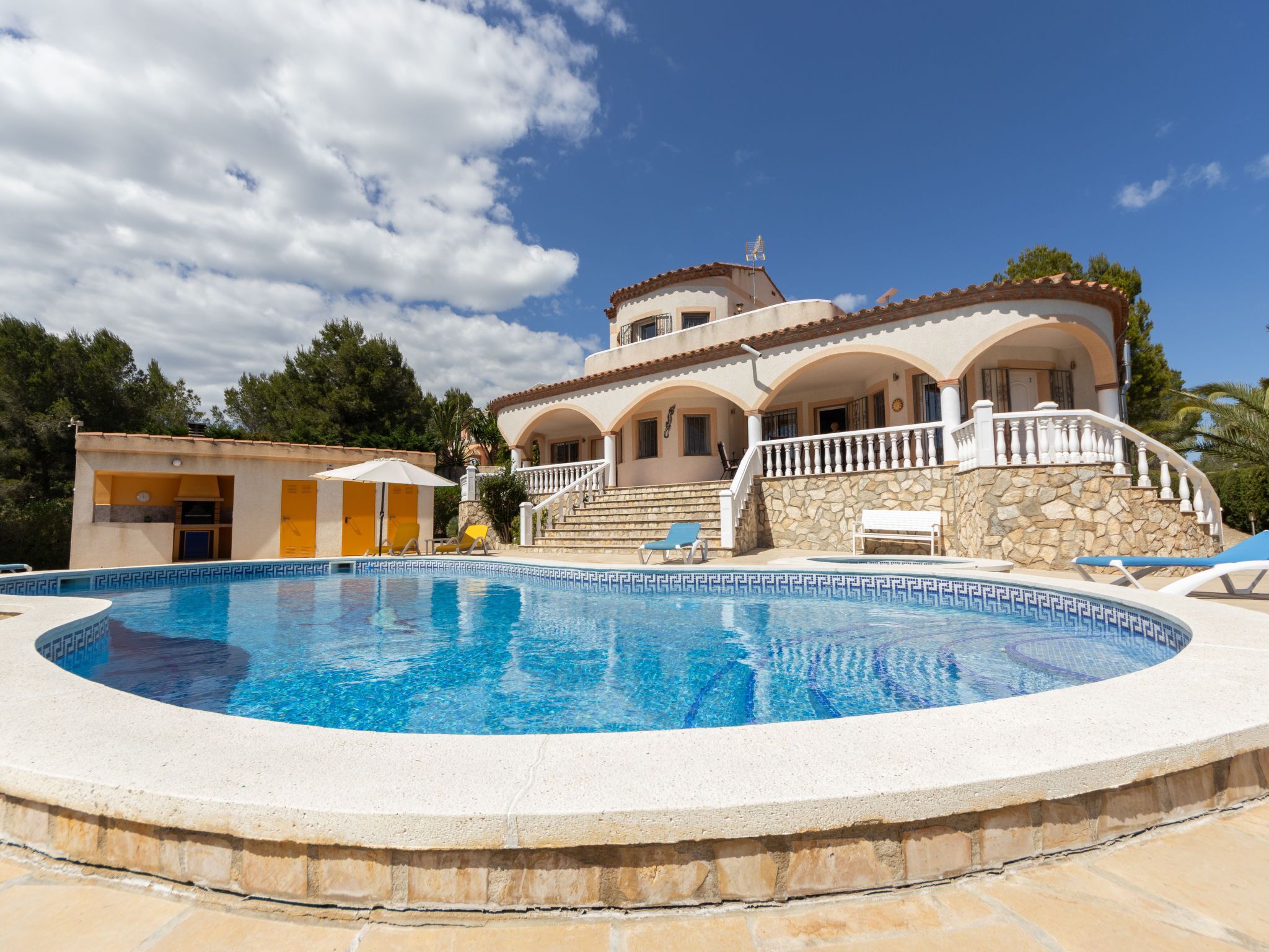 A large, circular swimming pool with a stone border, surrounded by a two-story building with a balcony and a stone wall.