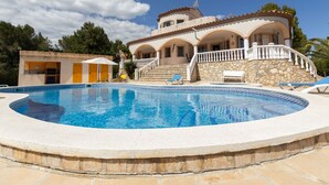 A large, circular swimming pool with a stone border, surrounded by a two-story building with a balcony and a stone wall.