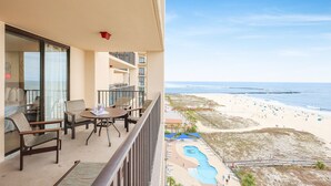 Un balcon avec une table et des chaises, donnant sur une plage avec une piscine et des personnes se baignant ou se prélassant sur le sable.