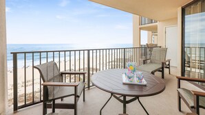 Un balcon avec une table ronde, deux chaises et un plateau avec des verres. Le balcon donne sur une plage où l’on aperçoit des gens et un ciel dégagé.