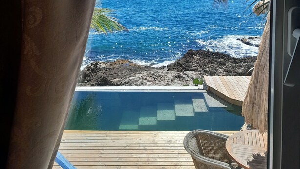 Blick von einem Zimmer mit großem Fenster auf das klare, blaue Meer, eine Holzterrasse und ein kleiner Pool.
