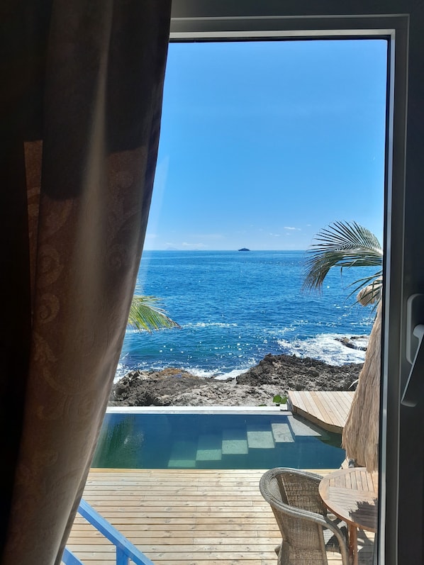 Vue depuis une chambre dotée d’une grande fenêtre donnant sur une mer bleue et limpide, une terrasse en bois et une petite piscine.