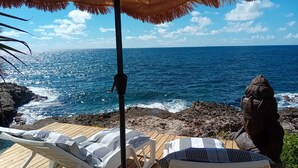 Eine Holzterrasse mit Liegestühlen und einer Tiki-Statue mit Blick auf eine felsige Küste und den Ozean.