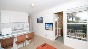A compact kitchen with white cabinets, a built-in microwave, and a small dining area with wicker chairs. There are two framed pictures on the wall, and a balcony with a view of the outdoors. - Beachside Shoal Bay Unit 2 (Shoal Bay)