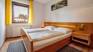 A bedroom with a wooden bed, two bedside tables, a window with yellow curtains, a radiator, and a painting on the wall.