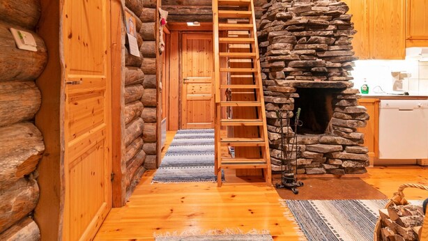 A log cabin interior with a wooden ladder leading to a loft bedroom, a stone fireplace, and wooden cabinets in the kitchen area.