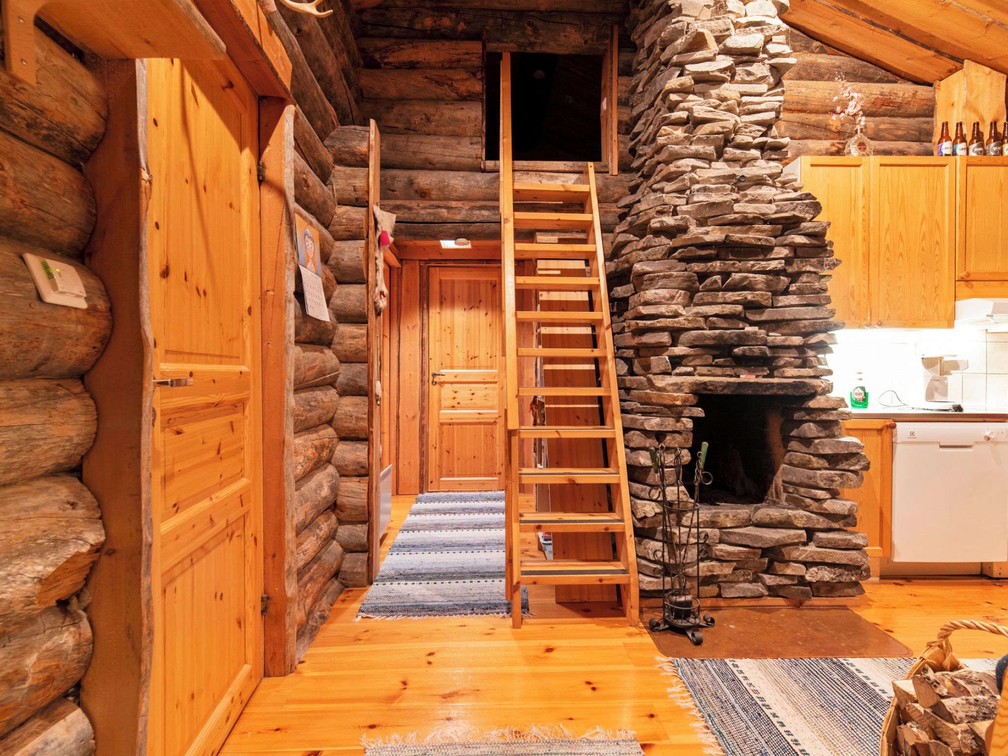A log cabin interior with a wooden ladder leading to a loft, a stone fireplace, and wooden cabinets.