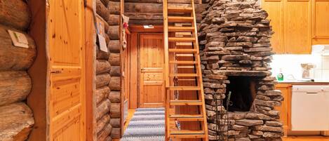 A log cabin interior with a wooden ladder leading to a loft, a stone fireplace, and wooden cabinets.