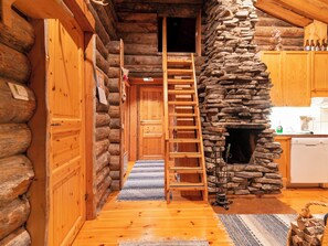 A log cabin interior with a wooden ladder leading to a loft, a stone fireplace, and wooden cabinets.