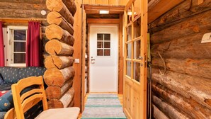 A log cabin interior with a wooden door, a small seating area with a chair and a sofa, and a window with curtains.