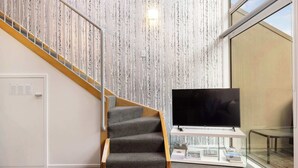 A staircase with a wooden handrail and carpeted steps, a flat-screen TV on a white stand, and a bookshelf with books and magazines.