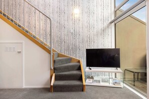 A staircase with a wooden handrail and carpeted steps, a flat-screen TV on a white stand, and a bookshelf with books and magazines.