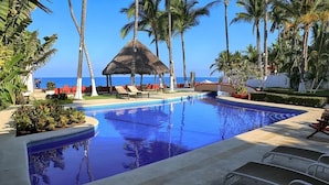 Un área de piscina con sillones de descanso, una estructura de techo de paja y palmeras.