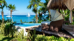 Zona junto a la piscina con una silla de mimbre, una estructura con techo de paja y vista al océano con un velero.
