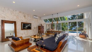 Amplia sala de estar con un sofá grande, una mesa de centro de madera y una mesa de comedor. Hay un televisor montado en la pared y un ventanal que ofrece vistas al exterior.