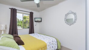 Een slaapkamer met een bed, een plafondventilator, een raam met gordijnen, een wandtelevisie en een decoratieve spiegel.