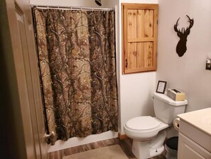 A bathroom with a shower curtain featuring a camouflage pattern, a wooden door, a deer head decoration, and a toilet. - The Lodge at 5J Hunts (Carrollton)