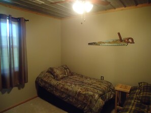 A bedroom with a camouflage-patterned bed, a wooden nightstand, and a wall-mounted saw blade decoration. - The Lodge at 5J Hunts (Carrollton)
